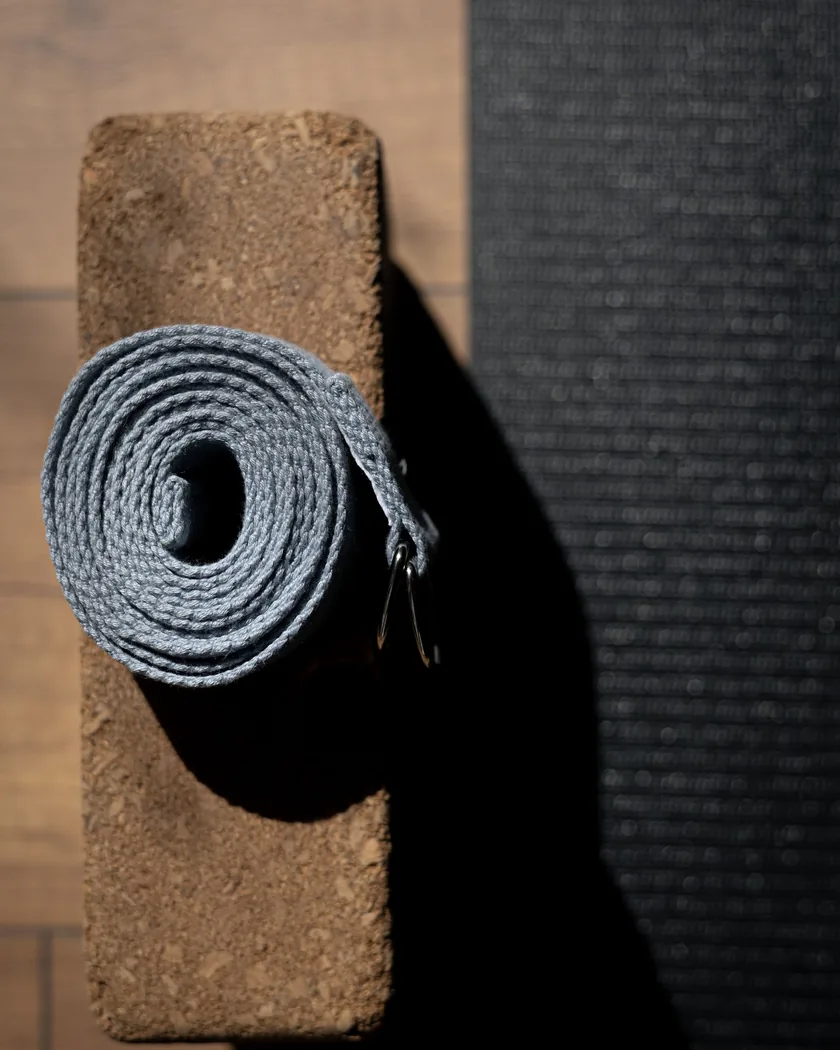 Bloque de corcho con un cinturón de yoga enrollado encima, apoyado sobre un suelo de madera junto al borde de una esterilla.